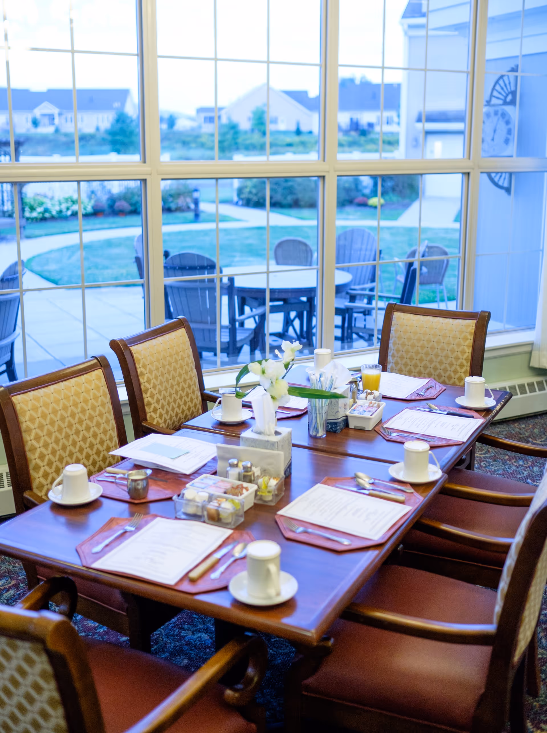 A dining table set for six with menus, cups, utensils, and a small flower arrangement in a room with large windows overlooking an outdoor patio area with chairs and tables.