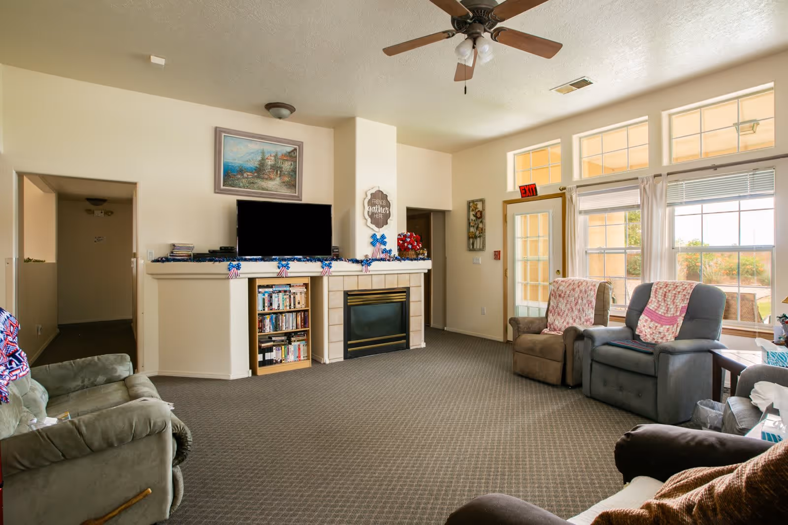 A cozy living room in an assisted living facility with several upholstered armchairs arranged around a fireplace and a flat-screen TV mounted above it. The room has large windows letting in natural light, a ceiling fan, and a carpeted floor. There are decorative items on the mantel and a bookshelf filled with DVDs or books below the TV.