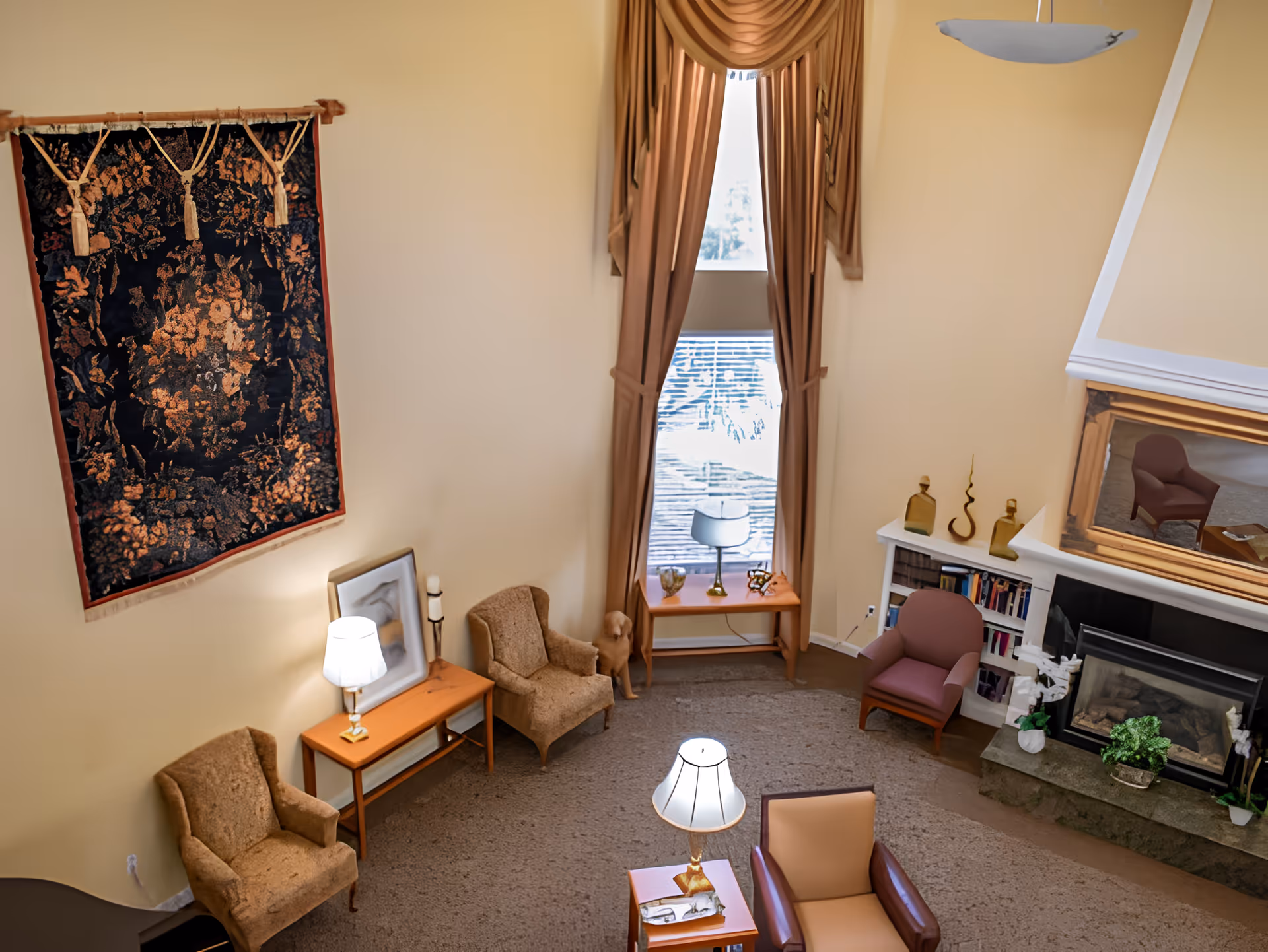A cozy living room with beige walls and carpet, featuring several armchairs arranged around a small wooden table with a lamp. A large window with brown curtains lets in natural light. There is a fireplace with a stone hearth and a large mirror above it, flanked by decorative items and bookshelves. A decorative tapestry hangs on one wall.