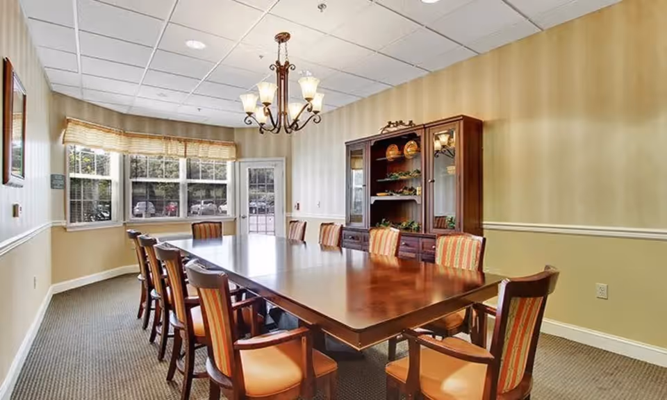 A well-lit dining room with a large rectangular wooden table surrounded by ten chairs with striped upholstery. There is a wooden cabinet with glass doors displaying decorative plates and plants against the far wall. A chandelier with multiple lights hangs from the ceiling. Large windows with blinds allow natural light into the room.