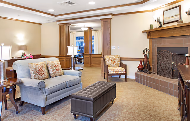 A cozy living room area with a gray upholstered loveseat with floral cushions, a brown leather tufted ottoman, a wooden side table with a lamp, and a fireplace with a decorative screen and mantel. The room has beige walls, carpeted floor, and wooden trim. In the background, there is a dining area visible through an open doorway.