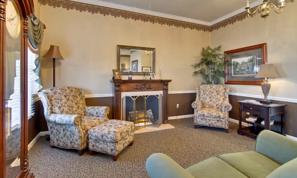 A cozy living room with floral upholstered armchairs and a matching ottoman, a green sofa, a wooden fireplace with a decorative screen, a mirror above the fireplace, a floor lamp, a table lamp on a side table, a framed painting, and a potted plant in the corner. The room has beige and brown walls with a decorative border near the ceiling and carpeted flooring.