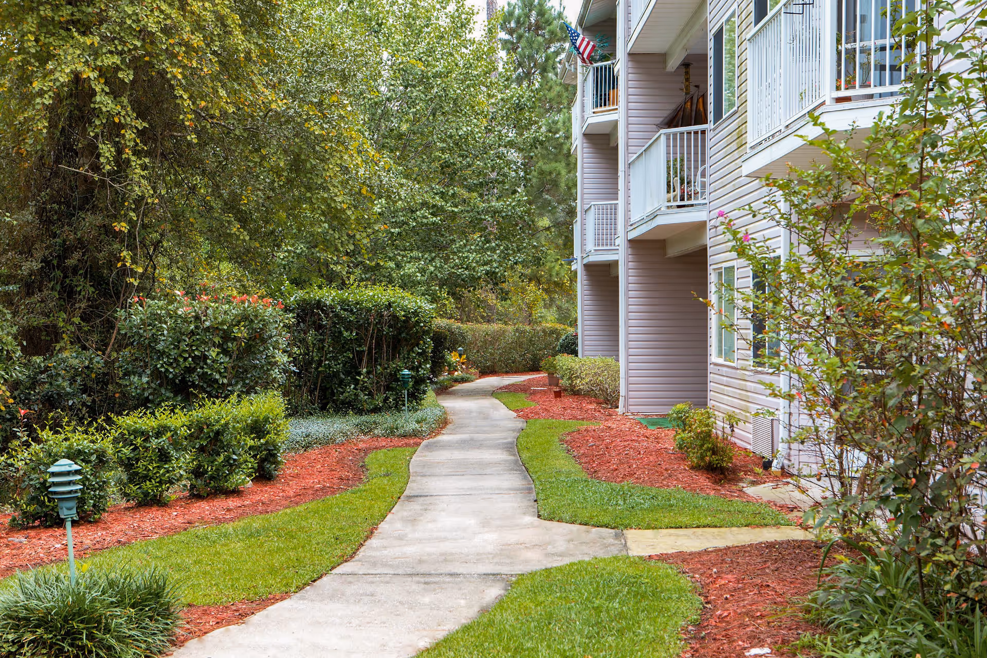 A winding concrete pathway bordered by well-maintained green grass and red mulch landscaping leads alongside a multi-story residential building with balconies and an American flag. The area is surrounded by dense green trees and shrubs.