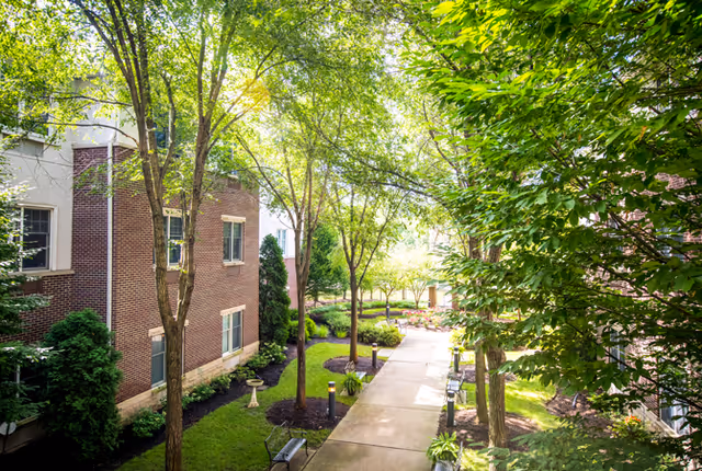Landscaped courtyard walkway between brick buildings lined with trees, benches, and greenery.