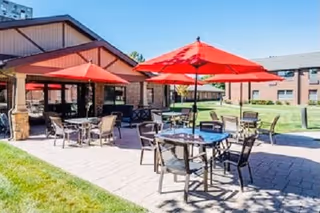Outdoor patio area at The Bridge at Valley View with multiple tables and chairs under red umbrellas on a paved surface, adjacent to a brick building with a clear blue sky above.