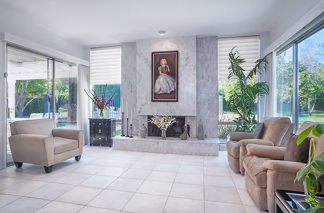 Bright modern living room with tiled floor, a central fireplace flanked by large windows, armchairs and potted plants.