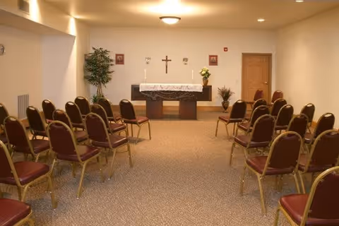 A small chapel or prayer room with rows of chairs arranged facing an altar. The altar is covered with a white cloth and has two candles, a crucifix on the wall behind it, and framed pictures on either side. There are plants placed near the walls, and the room has beige walls and carpeted floor.