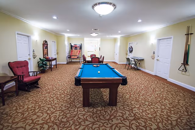 A spacious recreational room with a pool table in the center, a basketball arcade game at the far end, a keyboard on a stand, and several chairs and tables along the walls. The room has beige walls, patterned carpet, and multiple closed white doors.