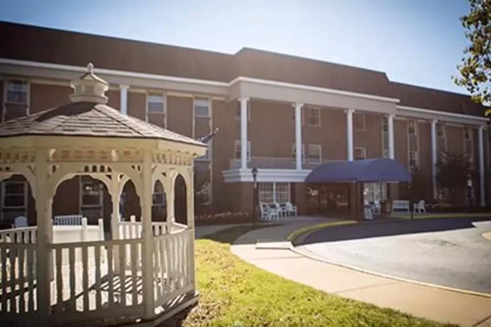 Exterior view of Symphony Manor Of Richmond, showing a three-story brick building with white columns and a blue awning over the entrance. In the foreground, there is a white wooden gazebo on a grassy area with a sidewalk and driveway leading to the entrance.