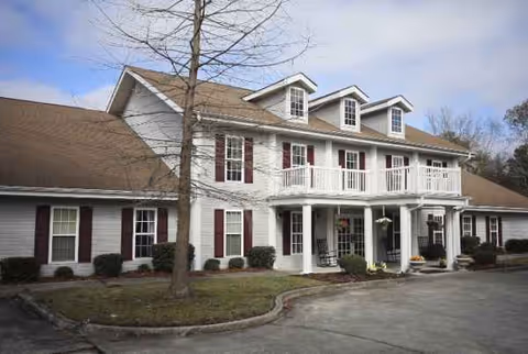 A two-story senior living facility exterior with white siding, dormer windows, and a wraparound porch.