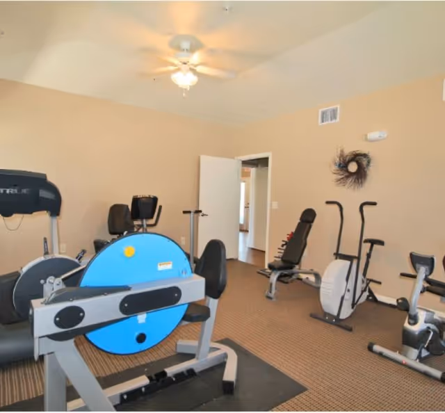 A small fitness room with various exercise equipment including a recumbent bike, a rowing machine with a blue flywheel, an elliptical trainer, and a weight bench. The room has beige walls, a ceiling fan with lights, and an open door leading to another area.