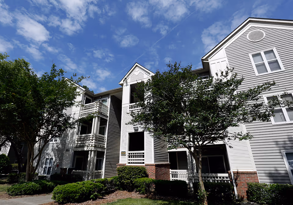 A multi-story senior living building with balconies, gray siding, brick accents, and landscaped shrubs under a blue sky.