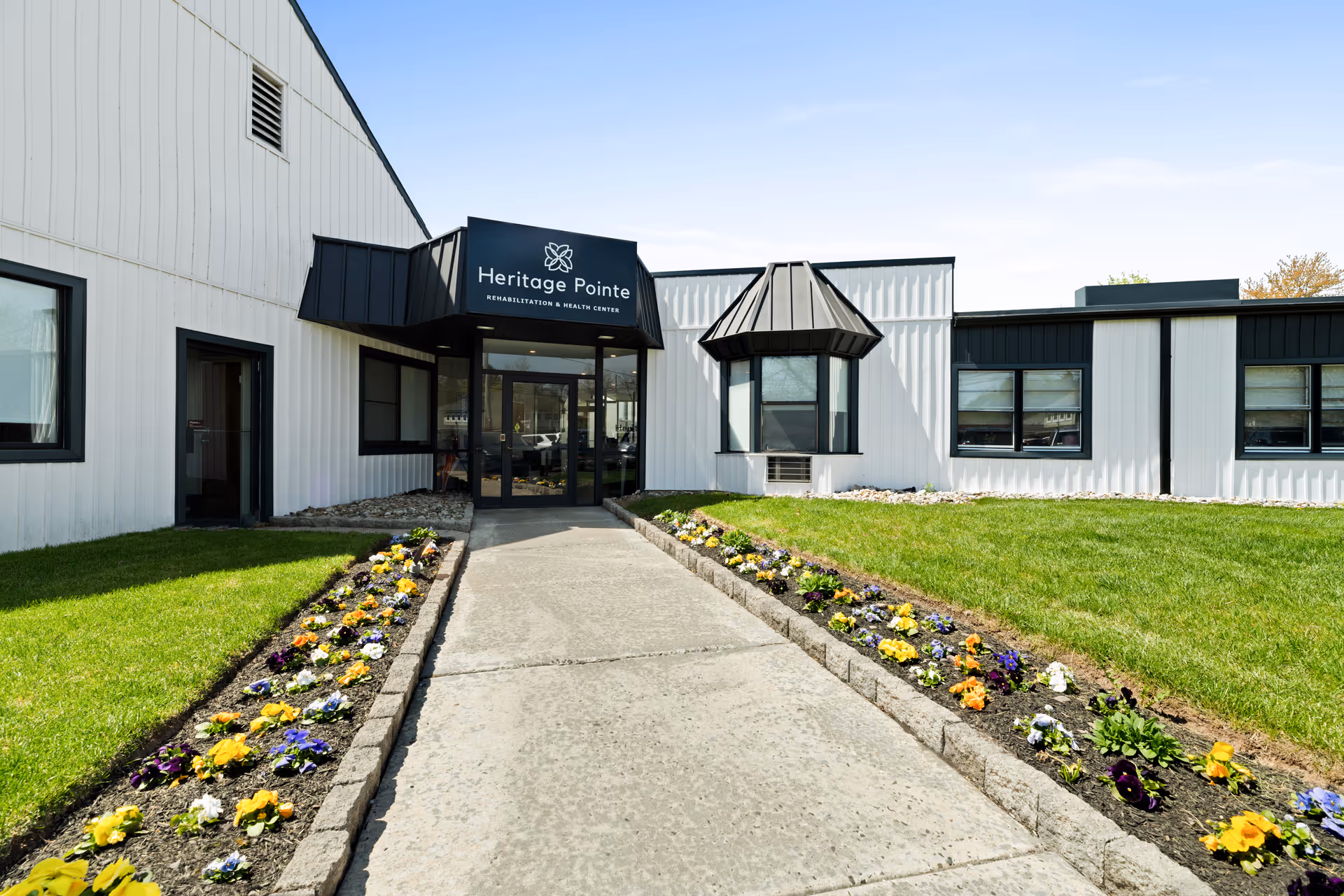 Front entrance of Heritage Pointe Rehabilitation and Healthcare Center with a concrete walkway flanked by flower beds and a black canopy sign.