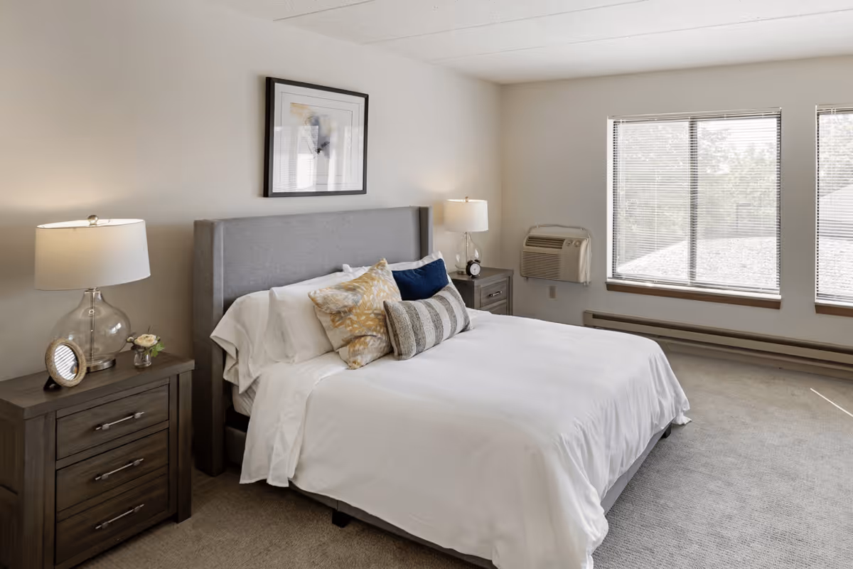 A bright and clean bedroom with a neatly made bed featuring white linens and multiple decorative pillows. There are two wooden nightstands on either side of the bed, each with a lamp. A framed abstract artwork hangs above the headboard. Large windows with blinds allow natural light to fill the room.