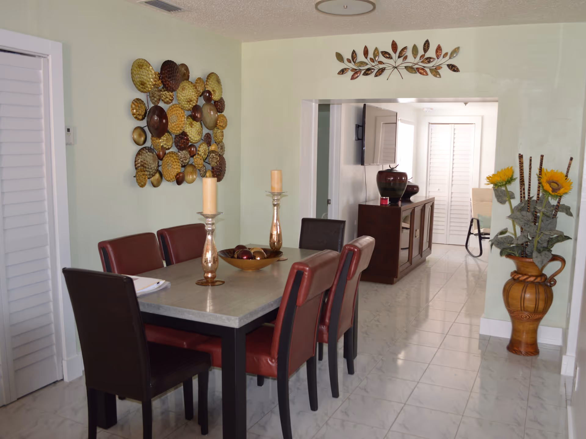 A dining room with a rectangular table surrounded by six chairs, four with red upholstery and two with black. The table has two tall candle holders and a decorative bowl with spheres. On the left wall, there is a metal wall art piece with circular patterns. In the background, there is a hallway leading to another room with a TV mounted on the wall and a cabinet below it. On the right side, there is a large vase with artificial sunflowers and decorative sticks. The floor is tiled with white marble-like tiles.