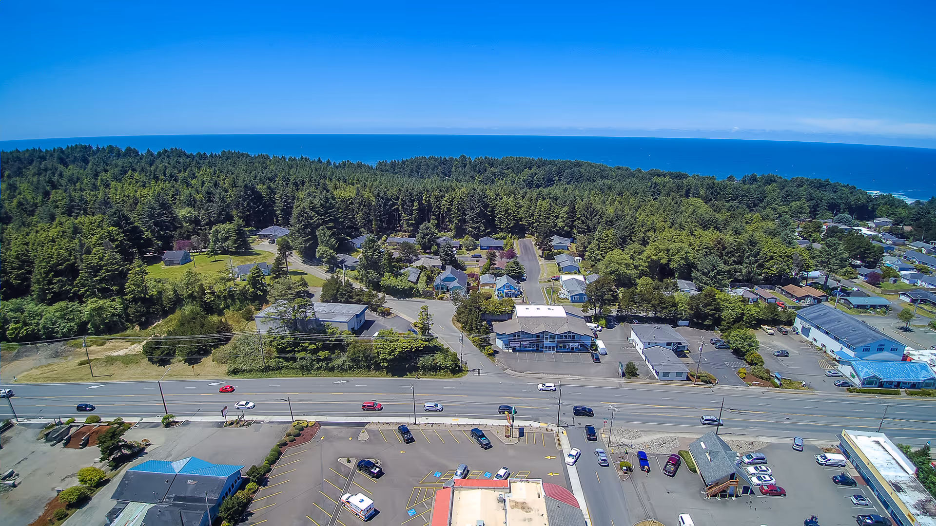 Aerial view of a coastal town with a large forested area, residential houses, and commercial buildings near a main road. The ocean is visible in the background under a clear blue sky. Several cars are parked in parking lots and driving on the road.