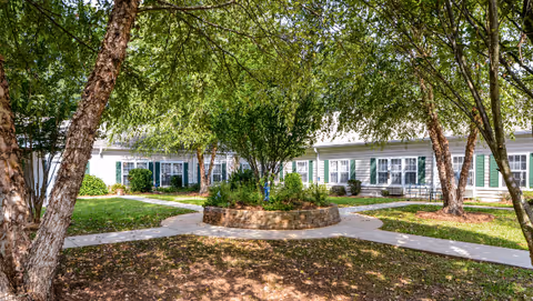 Outdoor courtyard area with a circular raised flower bed in the center surrounded by paved walkways and trees providing shade. White buildings with green shutters are visible in the background.