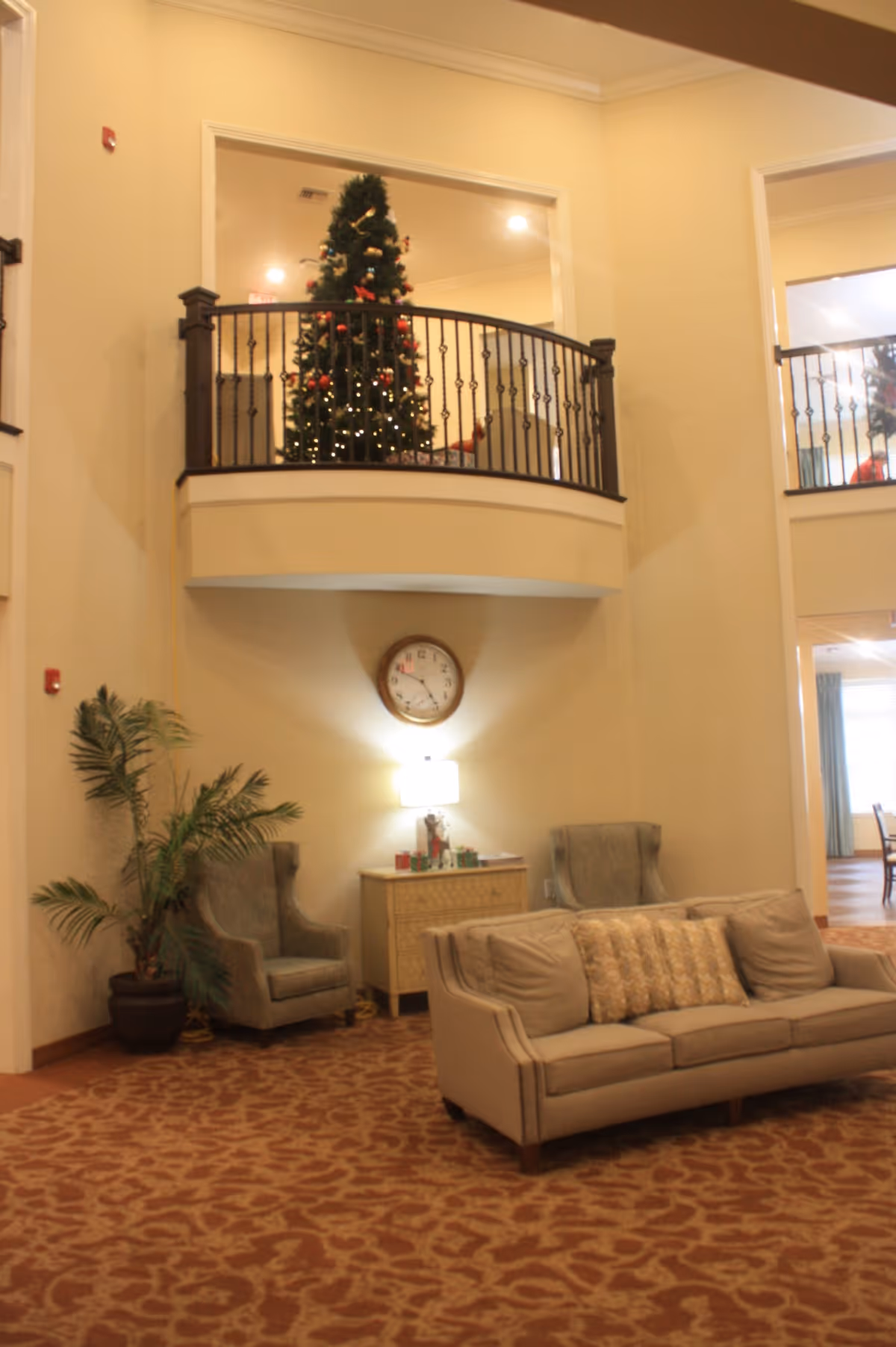 Interior view of a senior living facility common area with a beige sofa and two armchairs arranged around a small table with a lamp and clock on the wall above. A decorated Christmas tree is visible on the upper level behind a black railing. The room has warm lighting and patterned carpet.