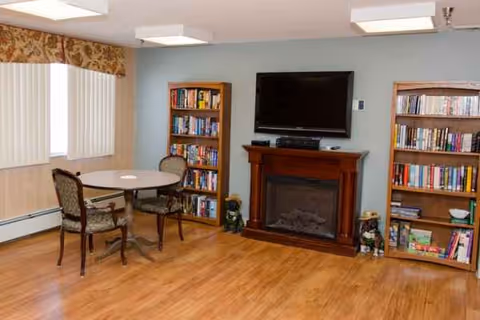 Common lounge area with fireplace, mounted TV, two bookshelves, and round table near windows.