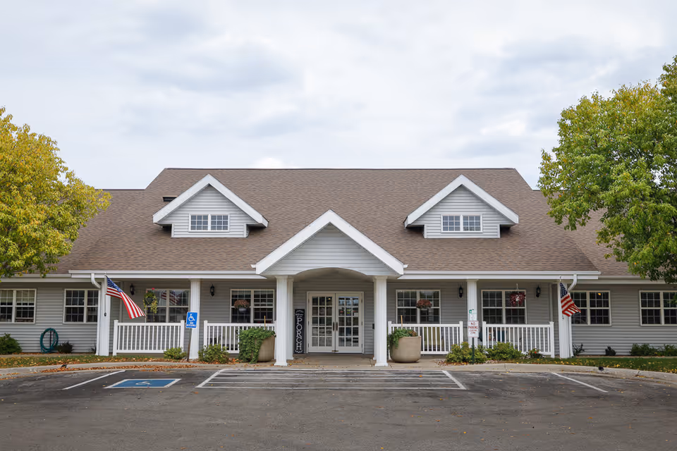 Front entrance of a single-story assisted living building with a covered porch, American flags, planters, and parking spaces.