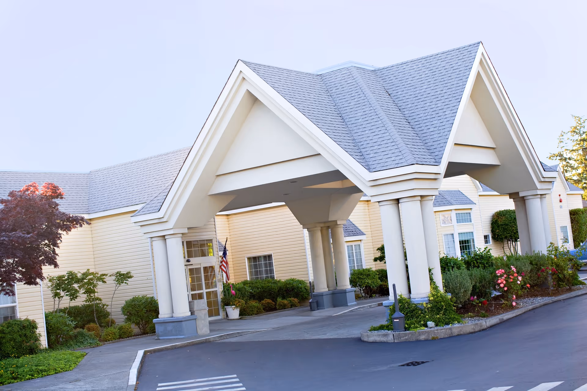 Front entrance portico of a senior living facility with tall columns, landscaping, and an American flag.