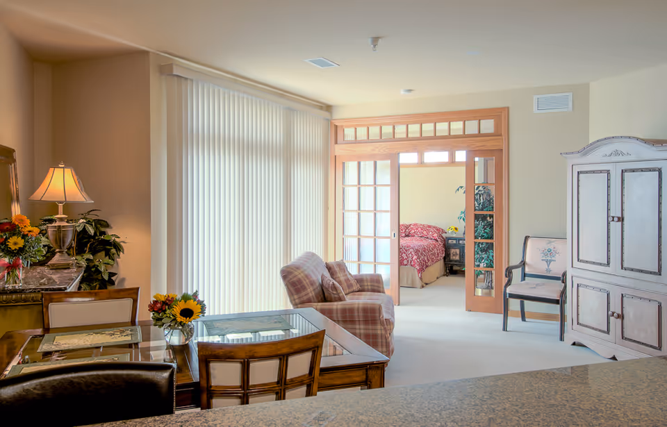 Bright open living and dining area with a plaid armchair, dining table, and glass-panel doors opening to a bedroom.