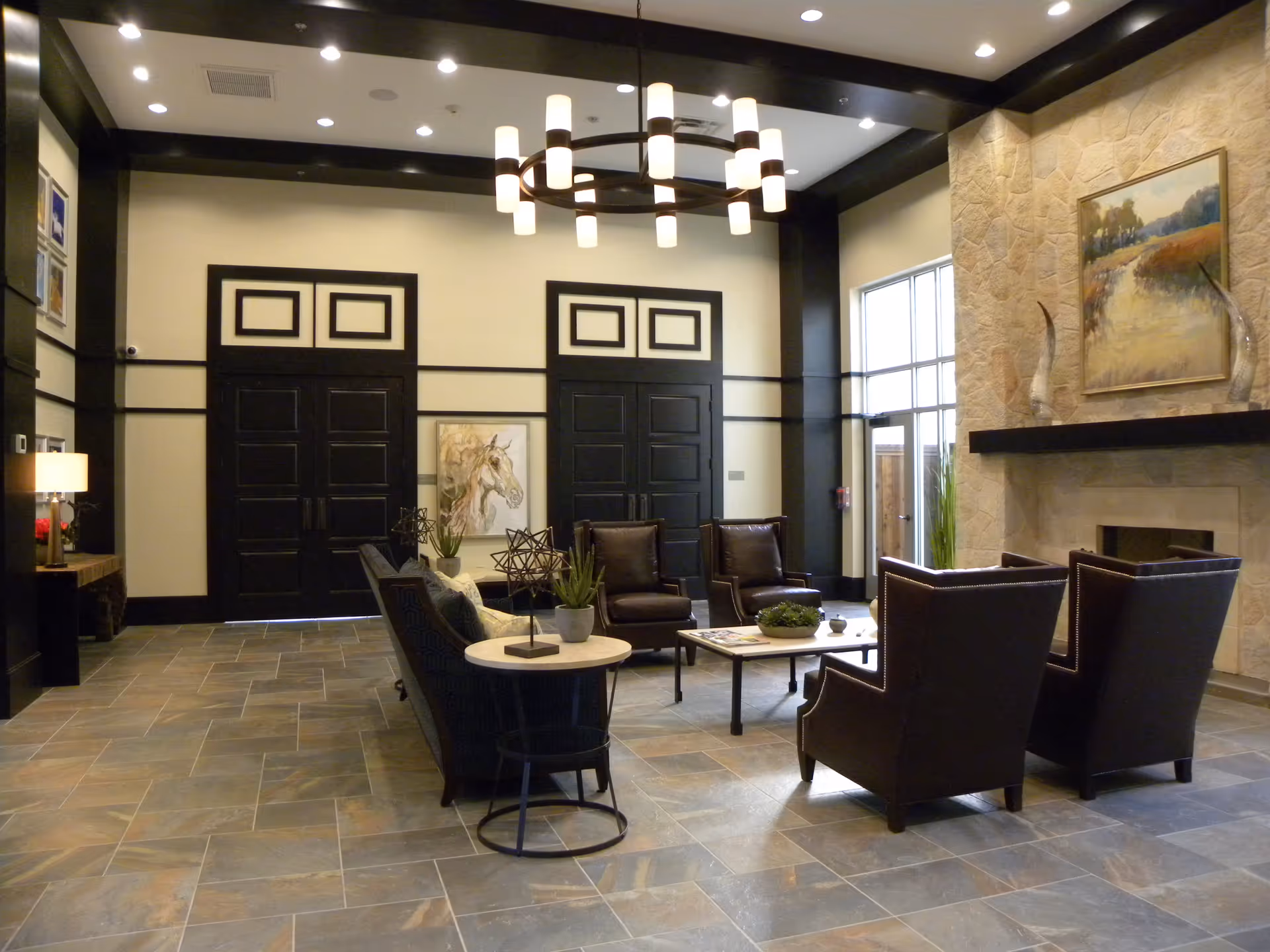 A spacious and elegant living room area with a modern chandelier hanging from the ceiling. The room features a stone fireplace with a landscape painting above it, two dark leather armchairs, a sofa, a coffee table with plants and magazines, and a side table with decorative items. The walls are painted in neutral tones with black trim, and there are large double doors and a window letting in natural light.