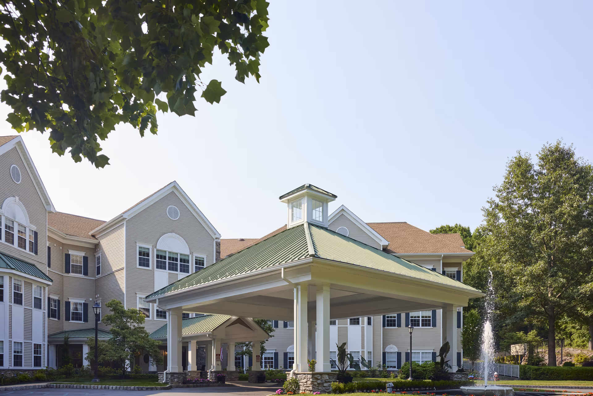 Exterior view of Brandywine Serenade by Monarch senior living facility showing a large covered entrance with a green roof, beige building with multiple windows, trees, and a water fountain in front.