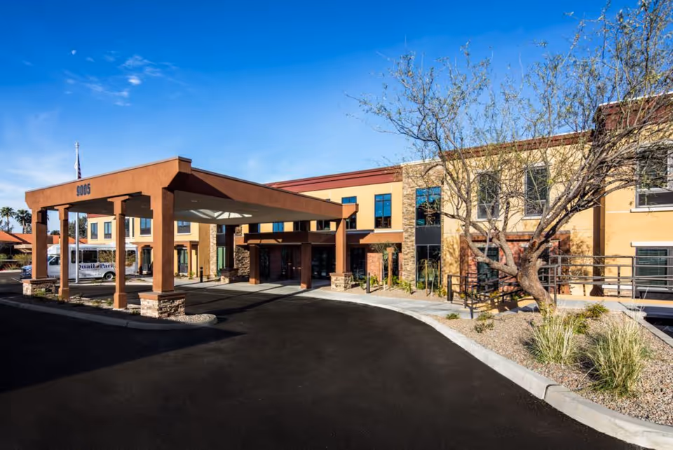 Exterior front entrance of a two-story senior living facility with a covered porte-cochere, curved driveway, and desert landscaping.