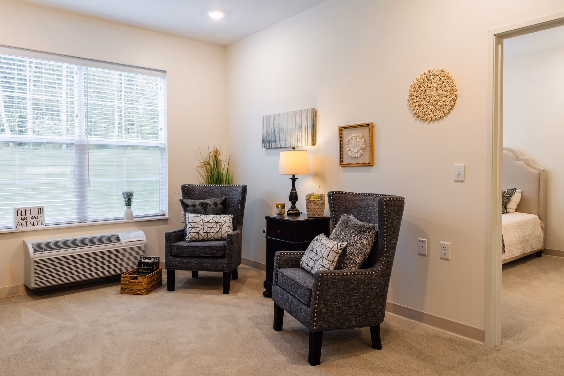 A cozy sitting area with two gray upholstered armchairs adorned with decorative pillows, a small black side table with a lamp, and wall art. A large window with blinds lets in natural light. To the right, an open doorway reveals a bedroom with a bed and pillows.