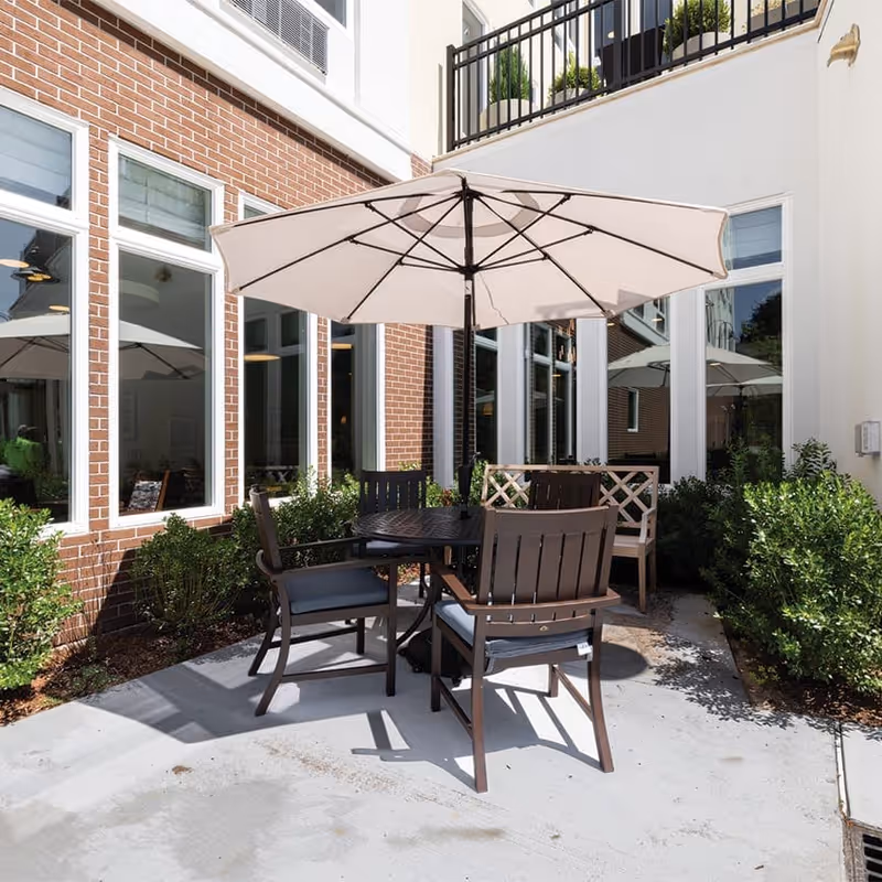 Outdoor patio area with a round table, four chairs, and a large beige umbrella providing shade. The patio is surrounded by brick and white walls with several windows and green bushes along the edges.