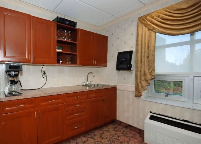 A small kitchenette area with wooden cabinets, a countertop with a coffee maker, a sink, and glassware on open shelves. There is a window with a gold curtain and a wall-mounted paper towel dispenser. The floor has a patterned carpet.