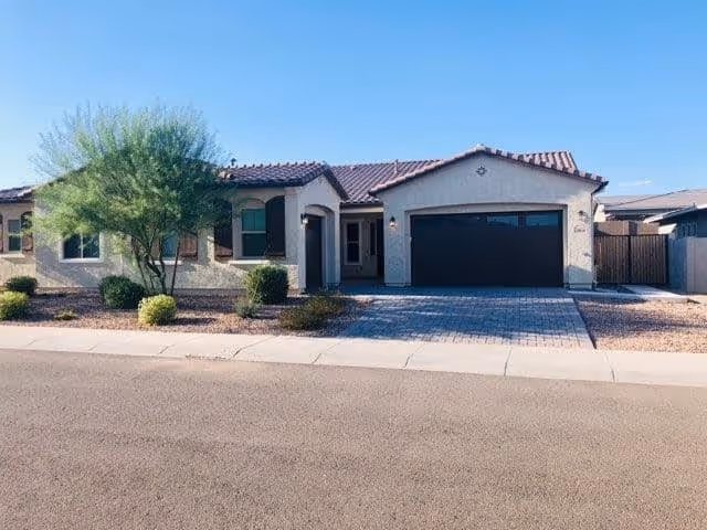 Single-story residential building with a tiled roof, a two-car garage with a dark door, and a small front yard featuring desert landscaping including bushes and a tree, under a clear blue sky.