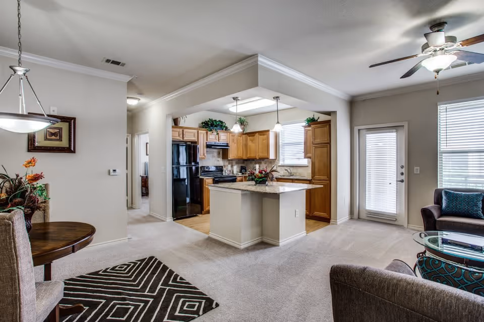 Open concept interior of a senior living facility showing a kitchen with wooden cabinets, black appliances, and a central island. Adjacent to the kitchen is a living area with a ceiling fan, glass coffee table, and upholstered chairs with blue patterned cushions. A small dining area with a round wooden table and upholstered chairs is visible on the left, along with a decorative rug and wall art. The space is well-lit with natural light coming through windows and a glass door.