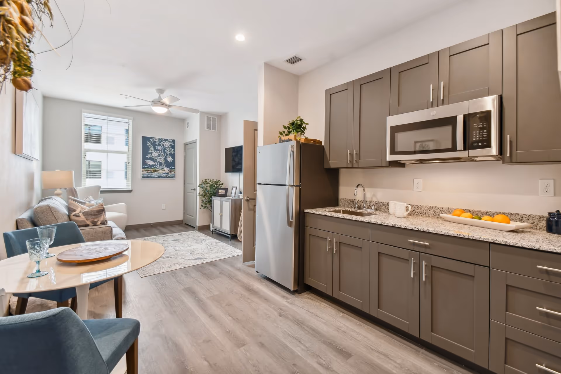 Open-plan apartment interior showing a modern kitchenette with gray cabinets and stainless appliances and a small living area with a sofa and dining table.