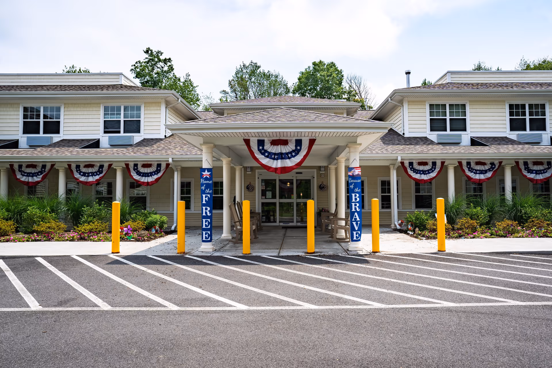 Front exterior view of a senior living facility decorated with patriotic red, white, and blue bunting. The building has a covered entrance supported by white columns with signs that read 'Home of the FREE' and 'Because of the BRAVE'. There are rocking chairs under the covered entrance and landscaped flower beds on either side.