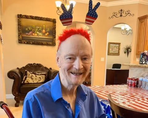 An elderly man wearing a blue shirt and a festive headband with red, white, and blue peace signs is smiling while sitting at a table with a red and white checkered tablecloth. Behind him is a living room area with a dark wooden couch and a framed painting on the wall.