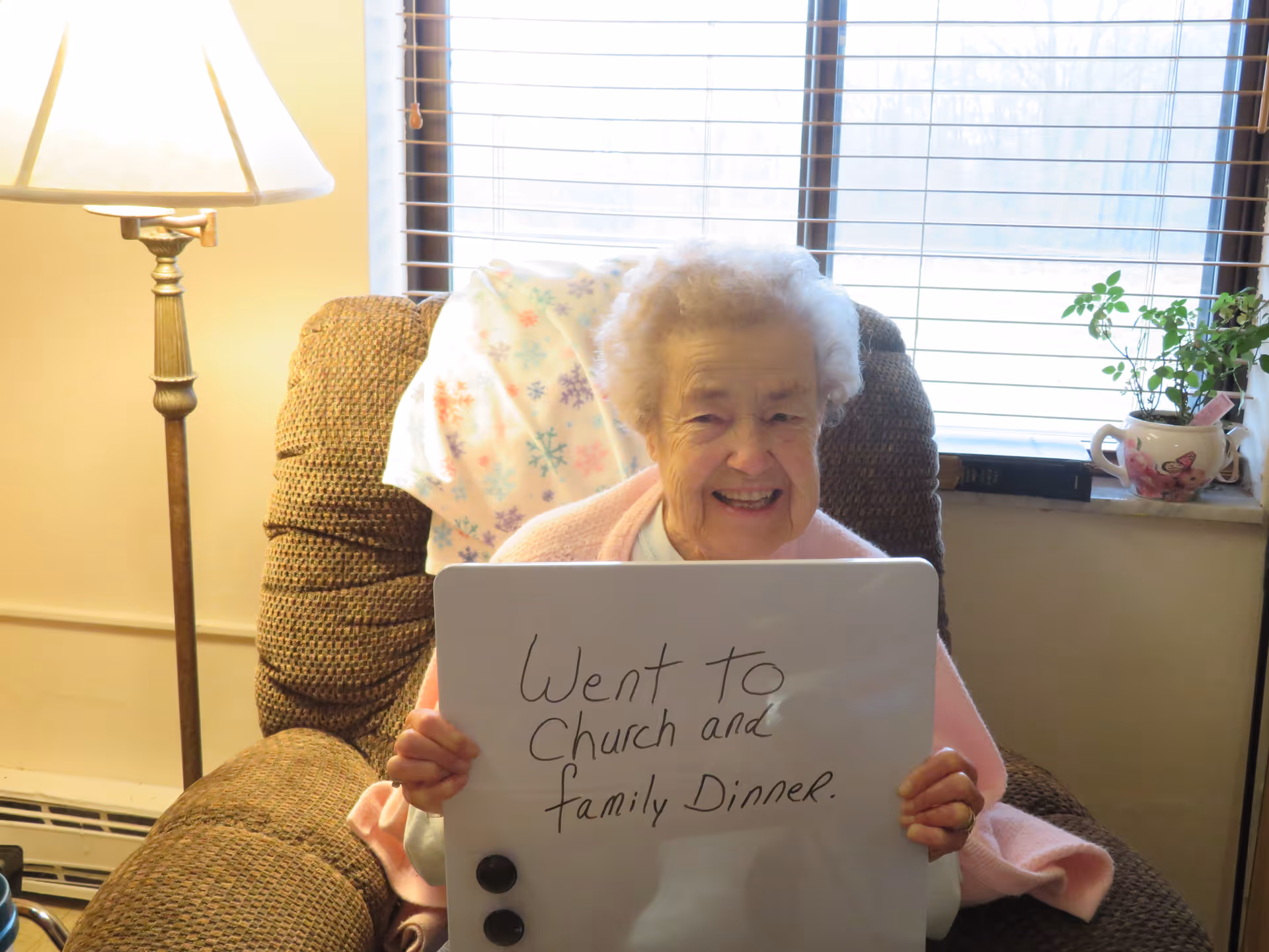 An older woman sits in a recliner holding a whiteboard that reads "Went to church and family dinner."