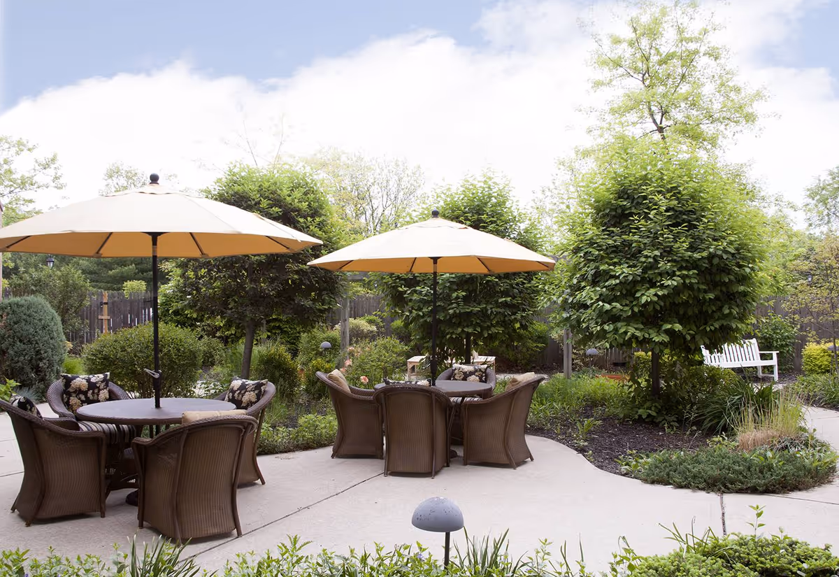 Outdoor patio area with two round tables, each shaded by a large beige umbrella. Each table is surrounded by four wicker chairs with floral cushions. The patio is surrounded by lush greenery, including trimmed bushes, small trees, and various plants. A white bench is visible in the background near a pathway.