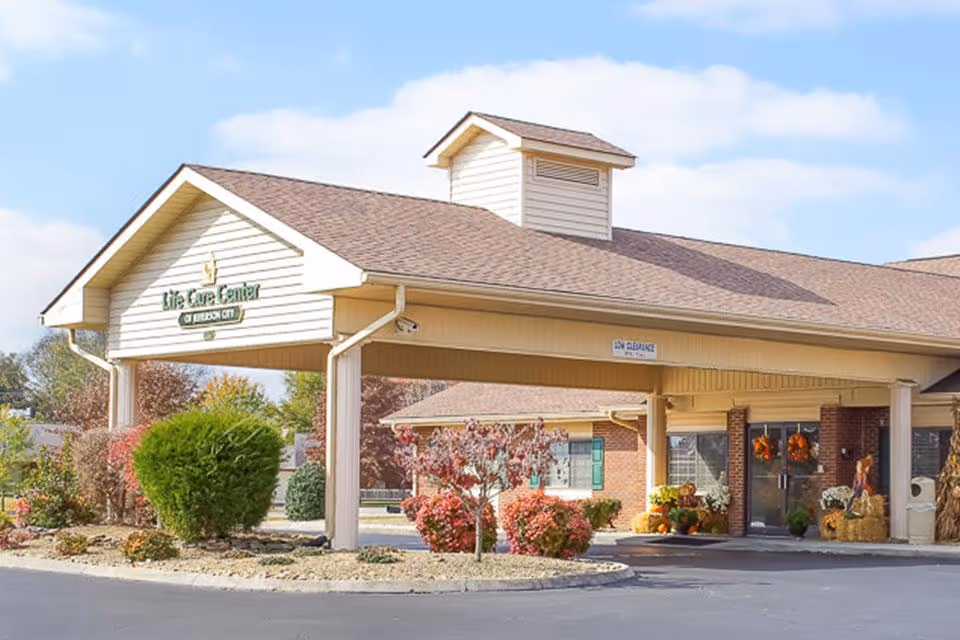Front exterior view of Life Care Center of Jefferson City showing the entrance with a covered driveway, landscaped bushes and trees, and fall decorations near the entrance doors.