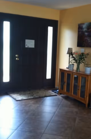 Interior view of an entryway with a dark wooden front door featuring two vertical frosted glass panels on either side. A small rug is placed in front of the door on a tiled floor. To the right, there is a wooden cabinet with glass doors, topped with a table lamp, a vase with flowers, and a potted plant. A framed picture hangs on the yellow wall above the cabinet.