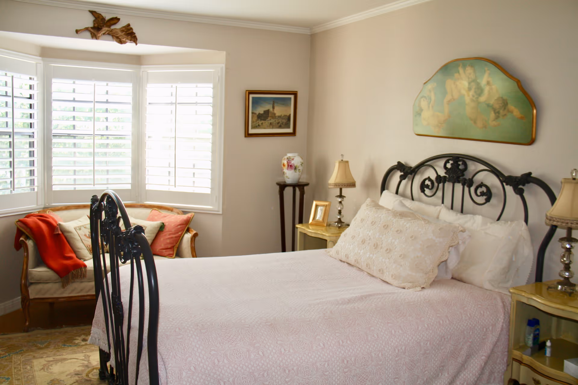 A cozy bedroom with a black wrought iron bed frame, a light pink bedspread, and decorative pillows. There is a vintage-style loveseat with cushions and a red throw blanket near a large window with white plantation shutters. The room also features two bedside tables with lamps, a small framed picture, a decorative vase on a stand, and wall art including a painting of cherubs above the bed.