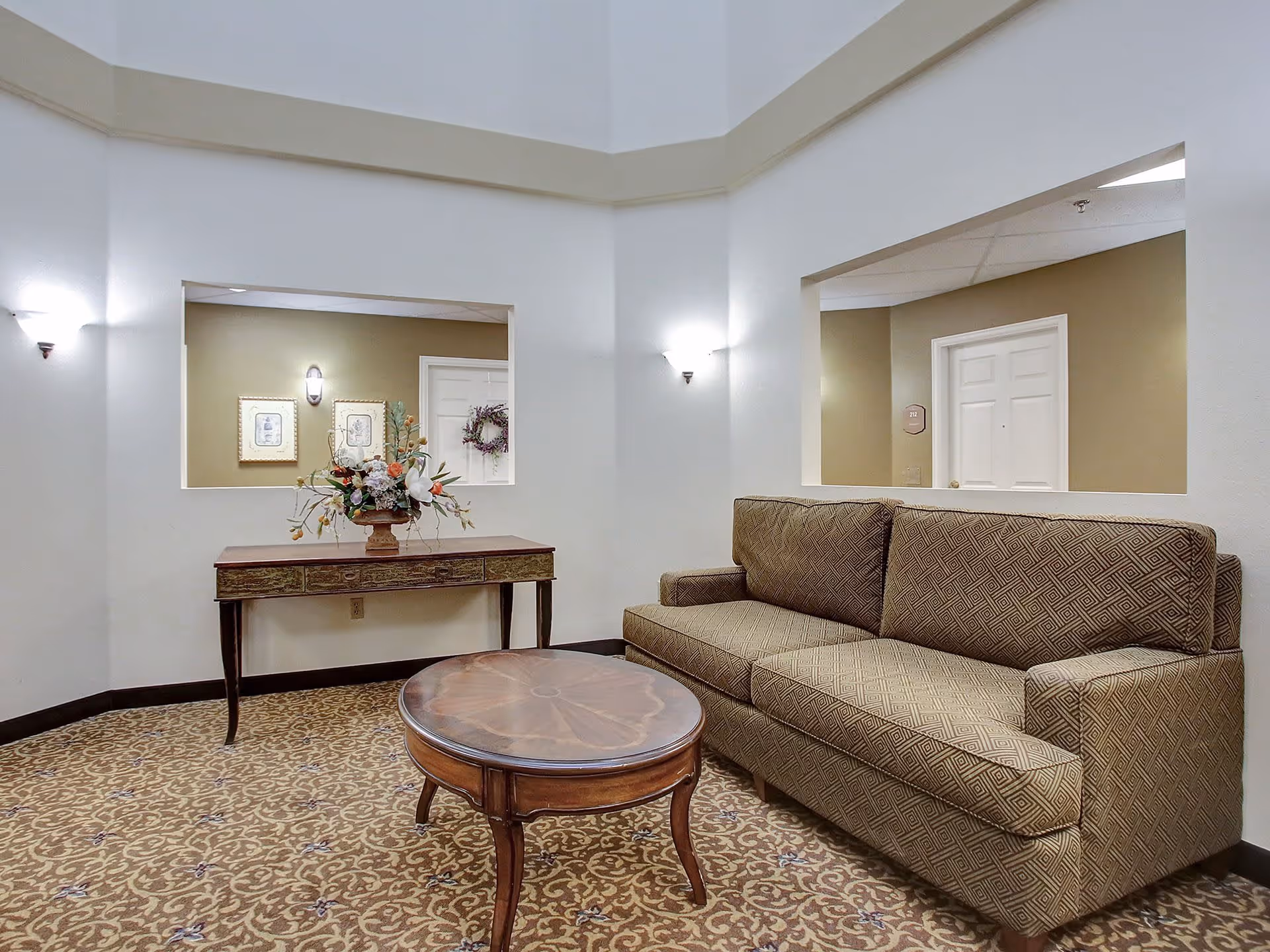 A cozy sitting area in a senior living facility with a patterned sofa, a round wooden coffee table, and a wooden console table with a floral arrangement. The walls are white with two large rectangular cutouts showing a hallway with doors and framed pictures. The carpet has a decorative pattern, and there are wall-mounted lights providing soft illumination.