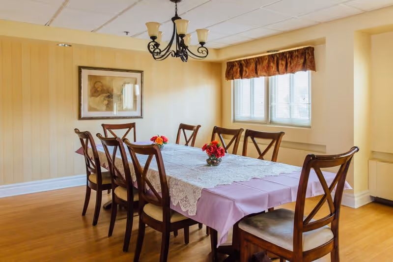 A dining room with a long table covered by a pink tablecloth and lace runner, small floral centerpieces, eight wooden chairs, a chandelier, and a window.
