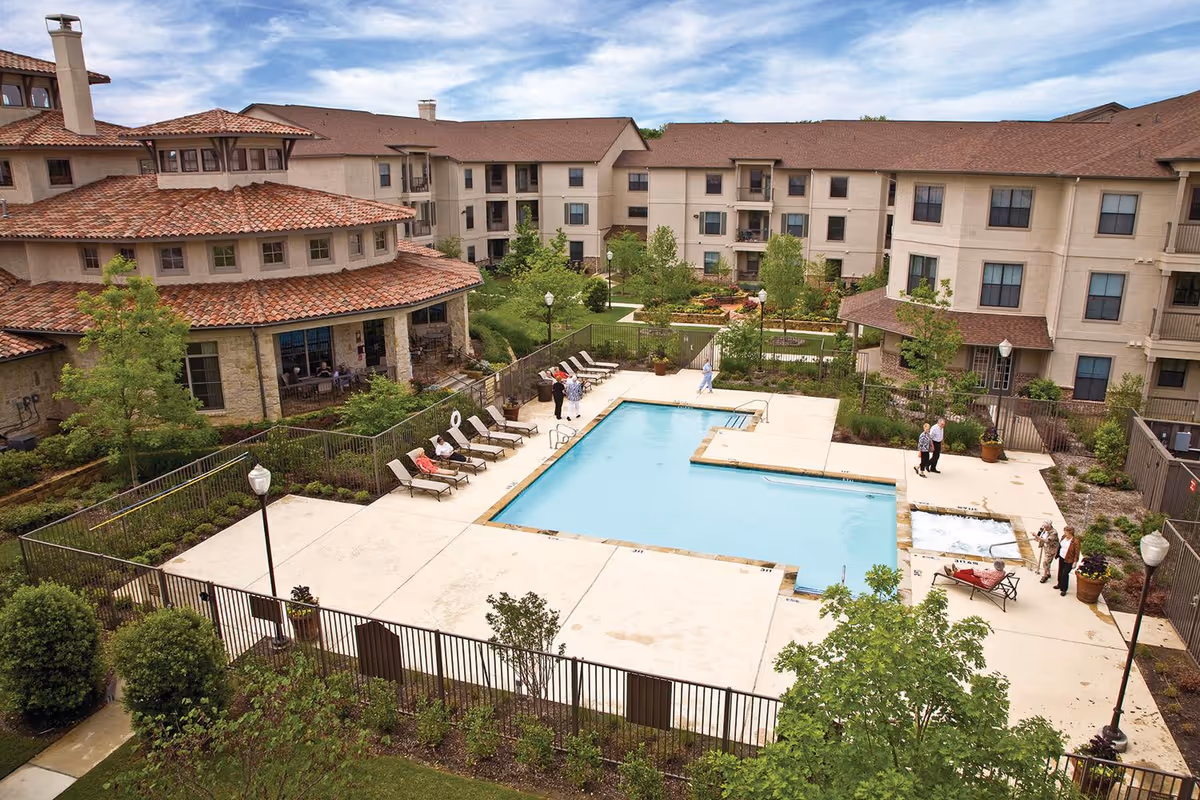A courtyard of a senior living facility featuring a fenced swimming pool with lounge chairs, landscaped gardens, and surrounding multi-story buildings.
