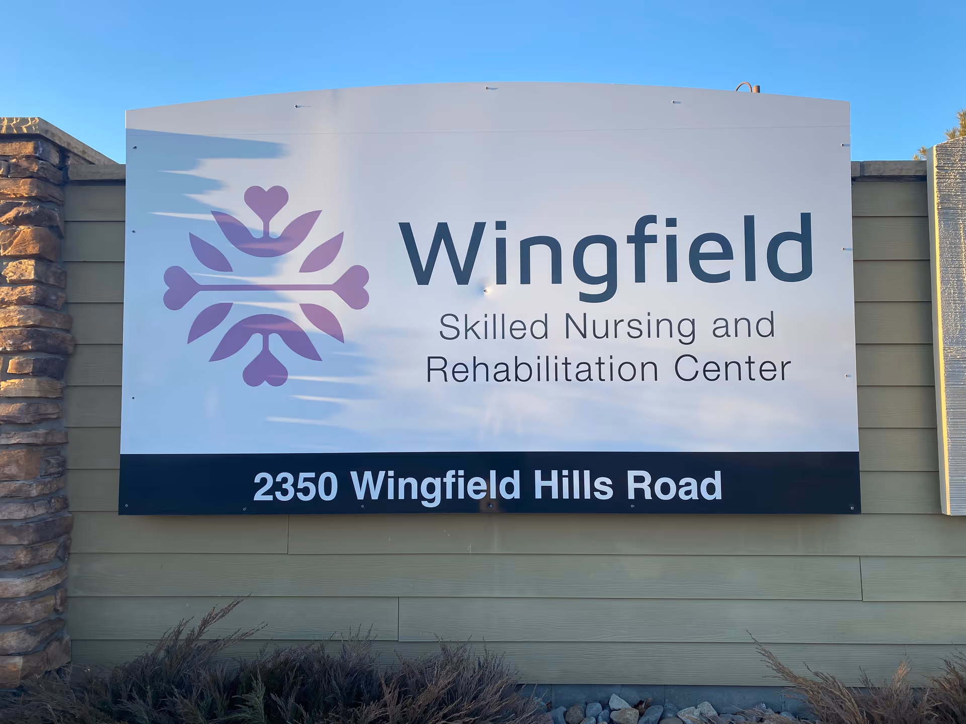 Outdoor sign for Wingfield Skilled Nursing and Rehabilitation Center with the address 2350 Wingfield Hills Road, mounted on a beige wall with stone pillars on the sides and some plants at the base.
