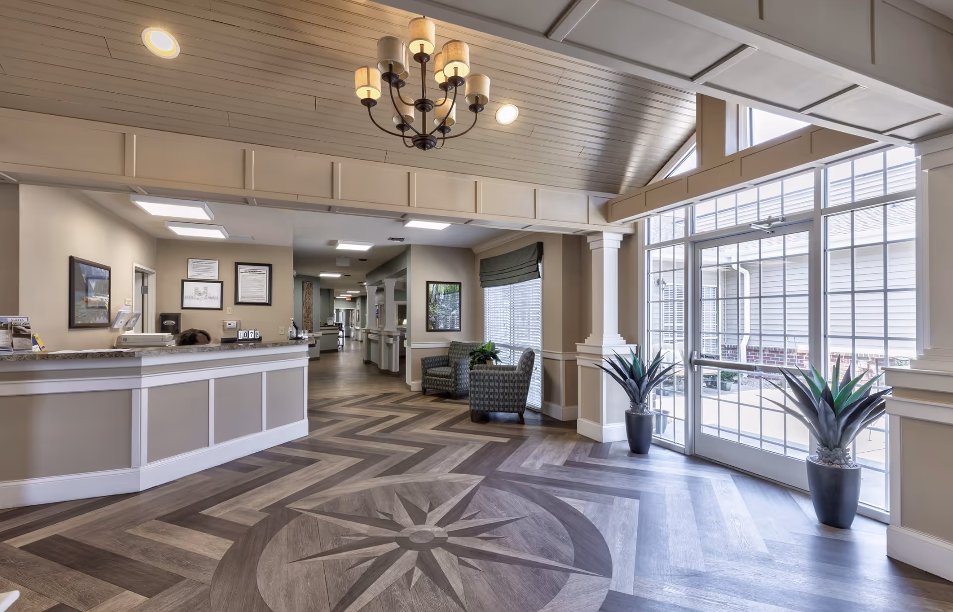Bright, spacious reception lobby with a curved front desk, seating area, decorative ceiling chandelier and patterned wood floor leading to large windows and glass doors.
