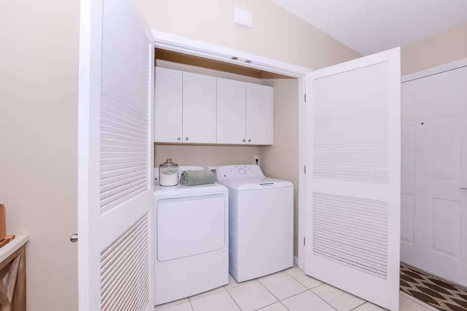 A laundry area with a white washing machine and dryer side by side, white cabinets mounted on the wall above them, and a green towel and a glass jar placed on top of the dryer. The area is enclosed by white bi-fold doors, and the floor is tiled.