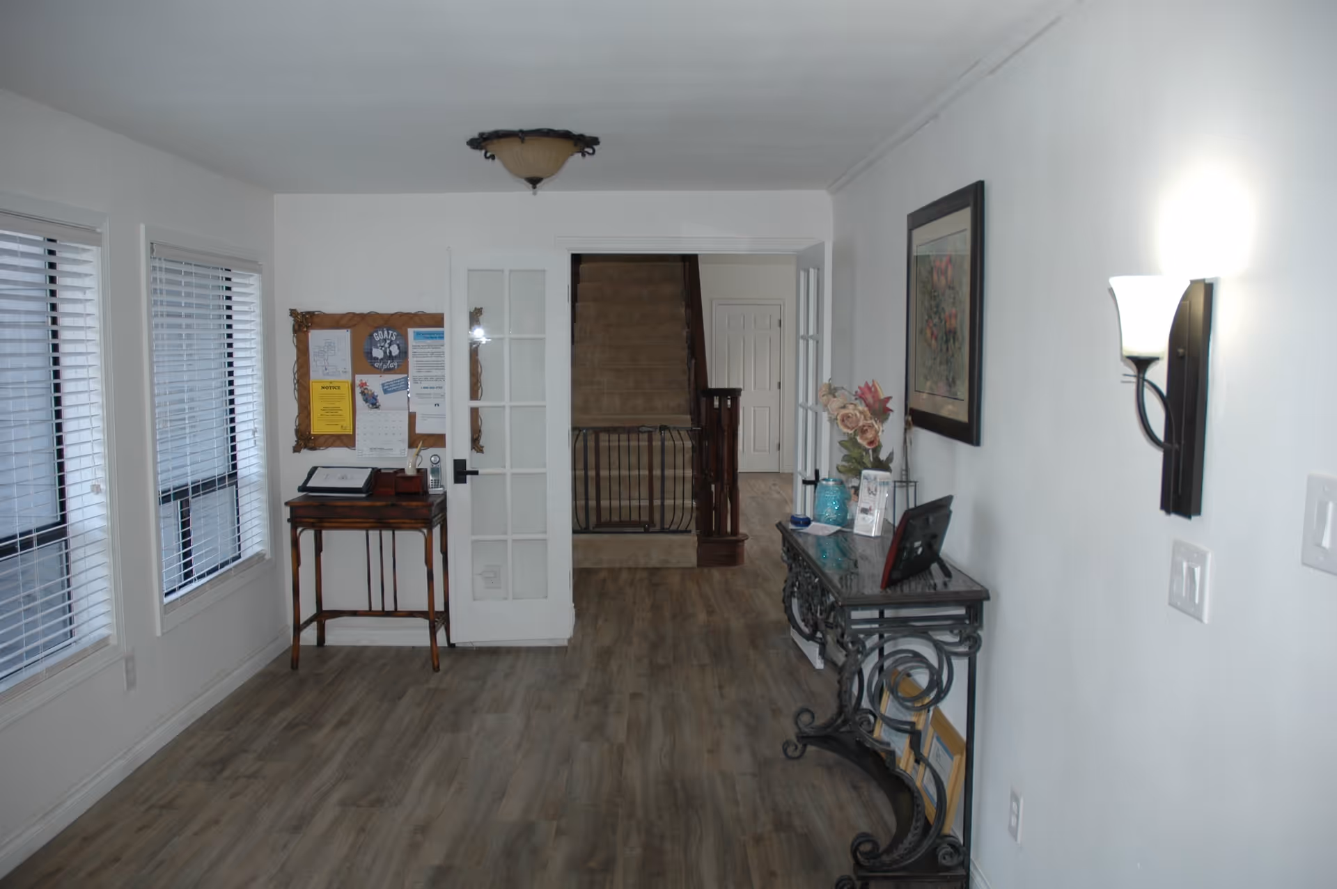 Interior hallway of a facility with light wood flooring, white walls, and two windows with blinds on the left. A small wooden table with a phone and bulletin board is on the left side near a glass-paneled door. On the right side, there is a decorative black metal console table with a glass top, holding a vase with flowers, framed pictures, and other small items. A wall-mounted light fixture is on the right wall, and a staircase with a baby gate is visible straight ahead.