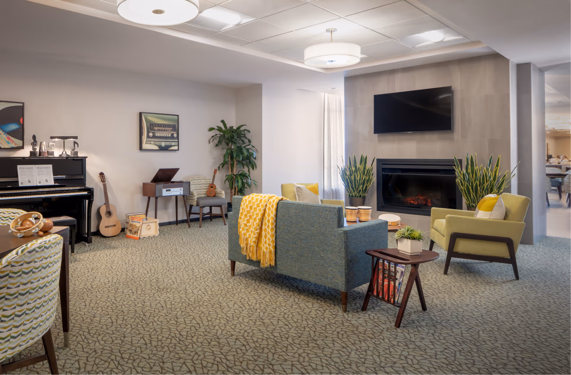 A cozy living room area with a blue sofa draped with a yellow blanket, a green armchair with a pillow, and a small wooden side table with plants and magazines. There is a modern fireplace with two tall green plants on either side and a flat-screen TV mounted above it. On the left side, there is a piano with sheet music, a guitar, a small chair with a ukulele, and a vintage record player. The room has patterned carpet and soft lighting from ceiling fixtures.